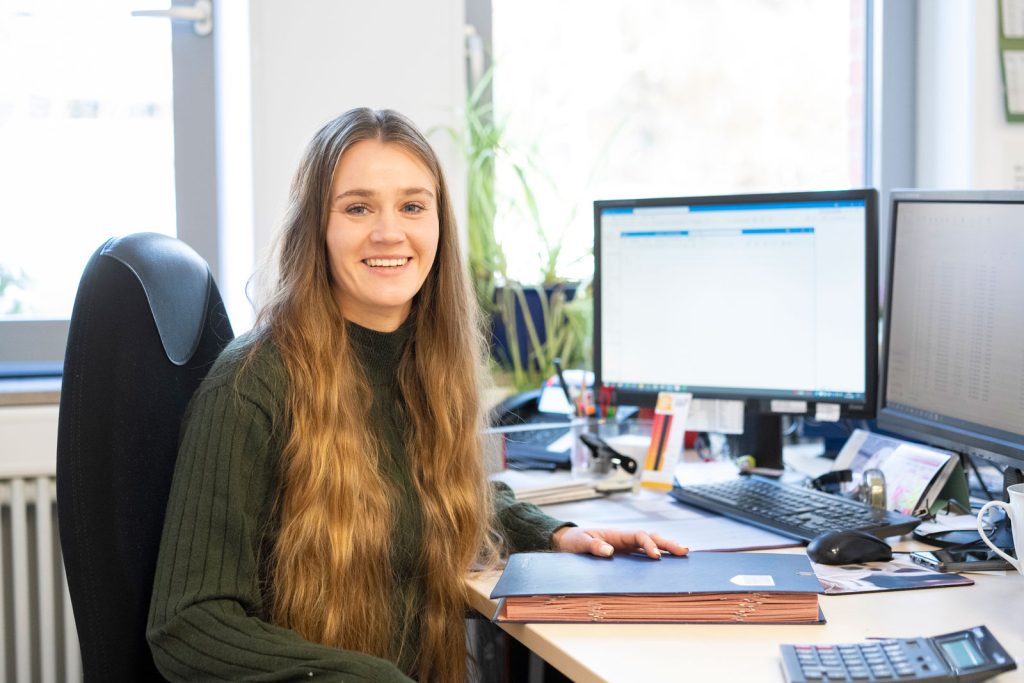 Eine junge Frau mit langen braunen Haaren sitzt l&auml;chelnd an einem B&uuml;rotisch mit zwei Computermonitoren und h&auml;lt einen Ordner in der Hand. Der Arbeitsplatz ist aufger&auml;umt und hell erleuchtet, B&uuml;romaterial und Pflanzen sind sichtbar.