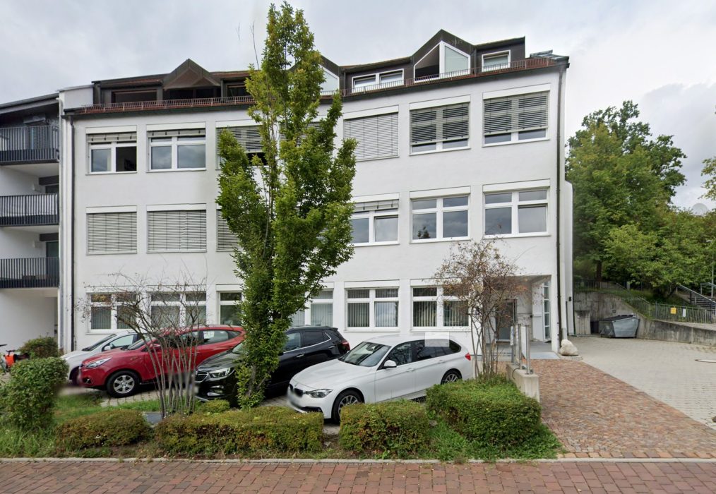 Der neue Standort der Freisinger Wohnbau. Ein wei&szlig;es dreist&ouml;ckiges Geb&auml;ude mit gro&szlig;en Fenstern, mehrere davor geparkte Autos und ein belaubter Baum, der die Sicht teilweise verdeckt; im Vordergrund sind ein gepflasterter B&uuml;rgersteig und etwas Gr&uuml;n zu sehen.